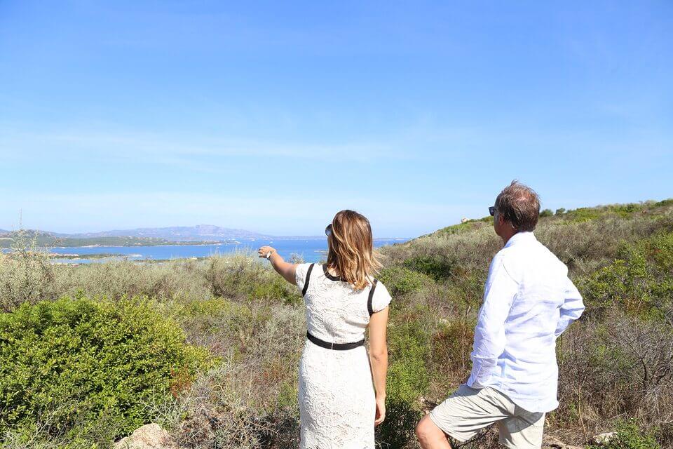 Ein Mann und eine Frau in Sommerkleidung stehen auf einem sonnigen Feld mit Blick auf ein klares, blaues Meer. Die Frau zeigt auf das Meer und beide betrachten philosophisch die Fernsicht. Um sie herum sind grüne Büsche und darüber ein klarer Himmel.