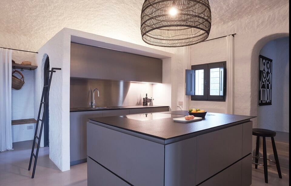 Eine moderne Küche mit eleganten grauen Schränken und Kochinsel, Edelstahlrückwand und weißen strukturierten Wänden. Der Raum, der ein philosophisches Konzept der Einfachheit verkörpert, ist mit einem großen, runden Kronleuchter aus schwarzem Draht und minimalistischem Dekor geschmückt.
