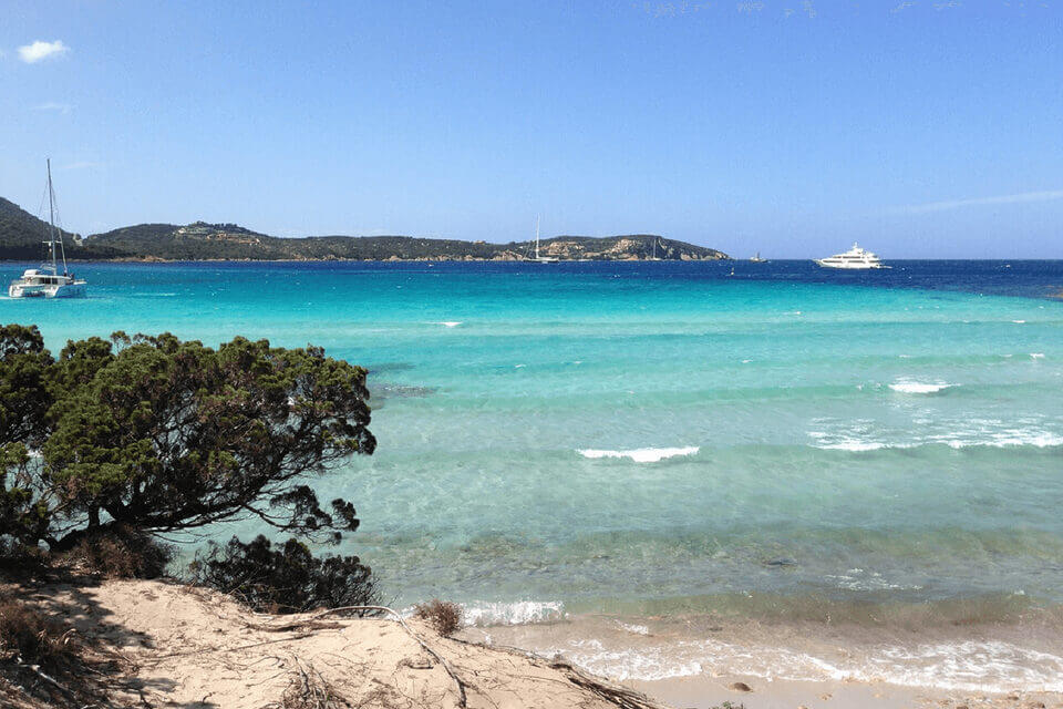 Eine malerische Strandszene in der Nähe des Feriendomizils Pevero mit türkisfarbenem Wasser und klarem Himmel. Sandstrand gesäumt von üppigem Grün, in der Nähe treiben Segelboote und eine Yacht. Im Hintergrund sind Hügel unter einer hellen Sonne zu sehen.