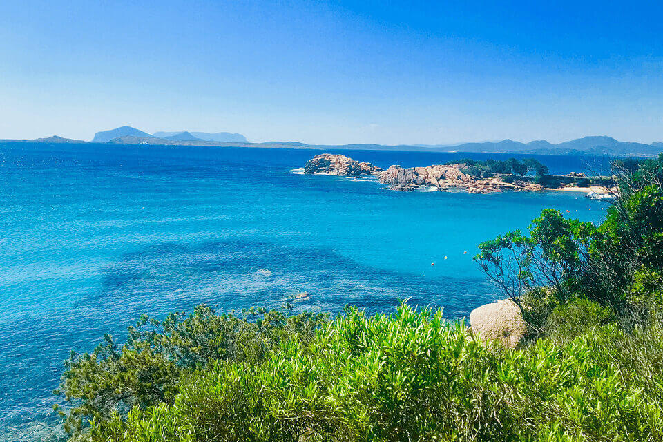 Eine lebendige Küstenlandschaft bei Romazzino mit klarem blauen Wasser, felsigen Ufern und üppiger grüner Vegetation mit einer Bergkette in der Ferne unter einem strahlend blauen Himmel.