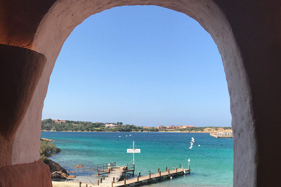 Eine gewölbte Steinöffnung umrahmt den Blick auf ein ruhiges türkisfarbenes Meer, einen Holzsteg mit einer Luxusyacht und entfernte Gebäude unter einem klaren blauen Himmel in Porto Cervo.