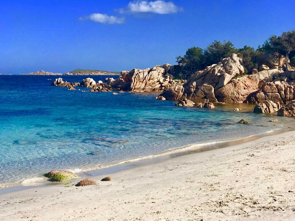 Eine ruhige Strandszene auf Sardinien mit klarem türkisfarbenem Wasser, weißem Sandstrand und abgerundeten Felsformationen an einem sonnigen Tag mit klarem blauen Himmel. Der Strand ist von grünem Gebüsch gesäumt, was die natürliche Schönheit noch verstärkt.