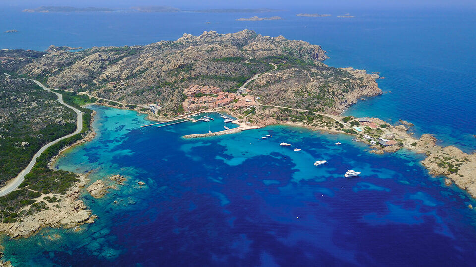 Luftaufnahme eines Küstengebiets in La Maddalena mit türkisfarbenem Wasser, verstreuten Booten, einem lebendigen Dorf mit roten Dächern und gewundenen Straßen inmitten felsigen Geländes, umgeben von dichtem Grün und dem offenen Meer.