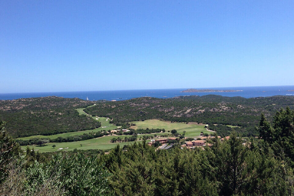 Üppig grüne Landschaft mit sanften Hügeln und einem luxuriösen Golfplatz in Porto Cervo. Der Blick geht über ein tiefblaues Meer in der Ferne, eingerahmt von klarem Himmel und dichtem Grün im Vordergrund.