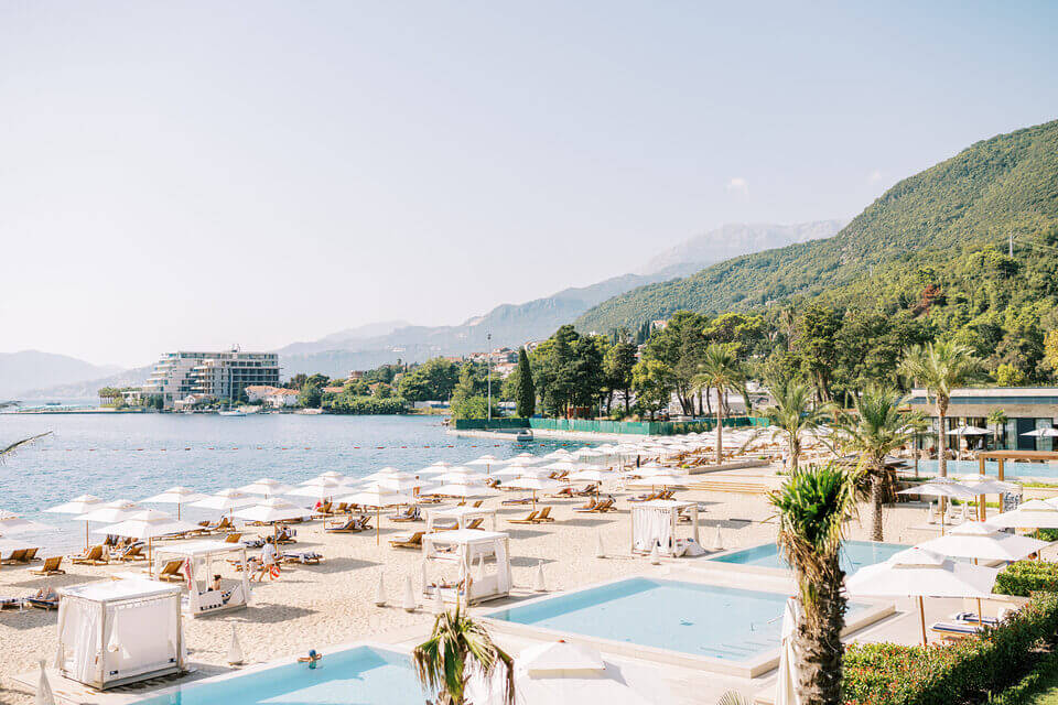 Ein malerisches Luxusresort an der Mittelmeerküste bei Portonovi mit Reihen von Liegestühlen an einem stillen blauen Pool, direkt angrenzend ein Sandstrand mit Palmen, mit Blick auf ein ruhiges Meer und Berge in der Ferne unter einem klaren Himmel.