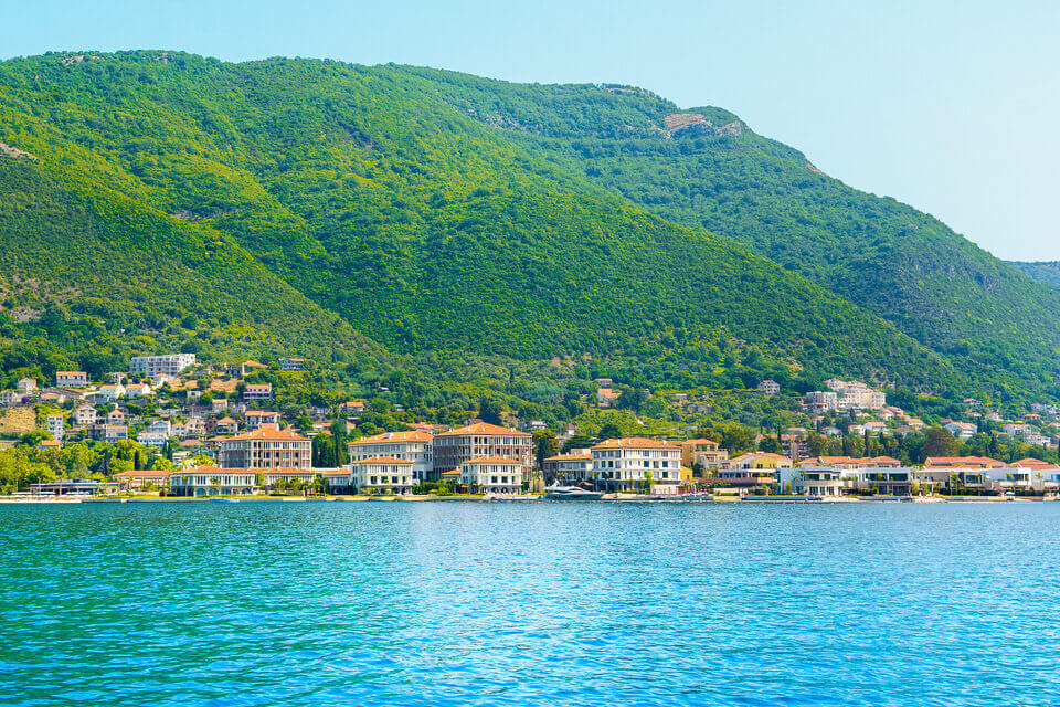 Ein malerischer Blick auf Portonovi mit üppigen grünen Hügeln im Hintergrund und einem Luxusresort am Wasser, in dem sich der klare Himmel und das ruhige blaue Wasser widerspiegeln.