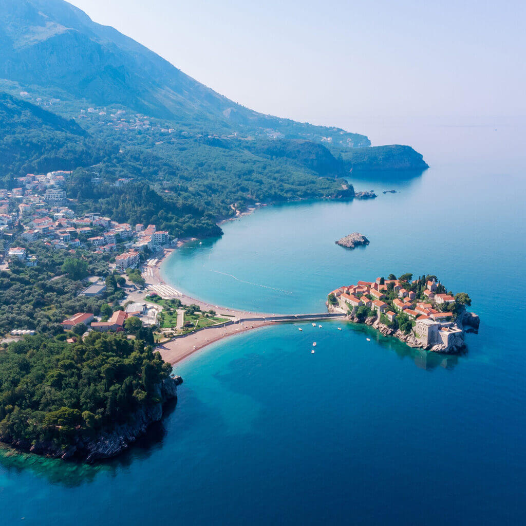 Luftaufnahme von Sveti Stefan in Montenegro, einem luxuriösen Ferienort mit geschwungenem Strand und klarem blauen Wasser, eingebettet zwischen üppig grünen Bergen und dem Meer, mit einer markanten kleinen Insel, die durch einen schmalen Damm verbunden ist.