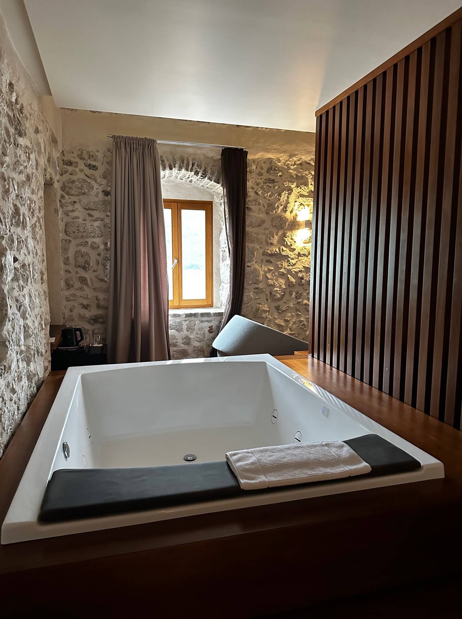 Ein ruhiges Badezimmer in einem Luxusresort mit einer großen, rechteckigen Whirlpoolwanne, umgeben von rustikalen Steinmauern. Eine Trennwand aus Holzlatten trennt den Wannenbereich vom Rest des Raums, und ein kleines Fenster sorgt für Tageslicht.