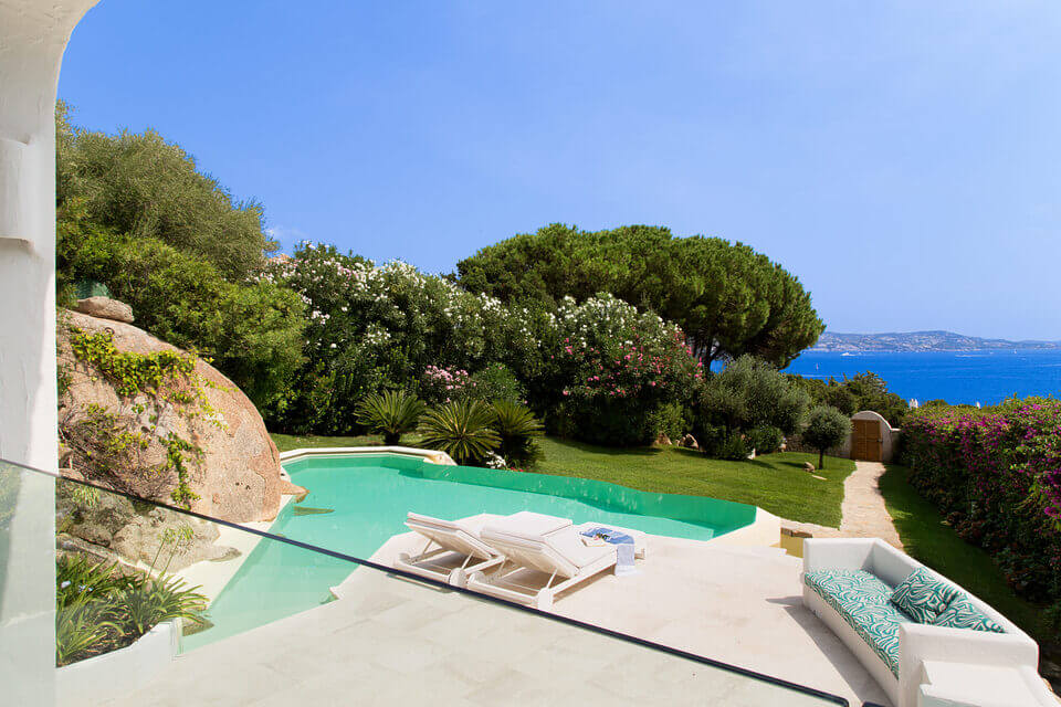 Luxuriöse Villa auf der Terrasse am Meer mit klarem blauen Himmel, geschwungenem Pool, weißen Sonnenliegen, üppigen Bäumen und blühenden Sträuchern und Blick auf ein ruhiges blaues Meer.