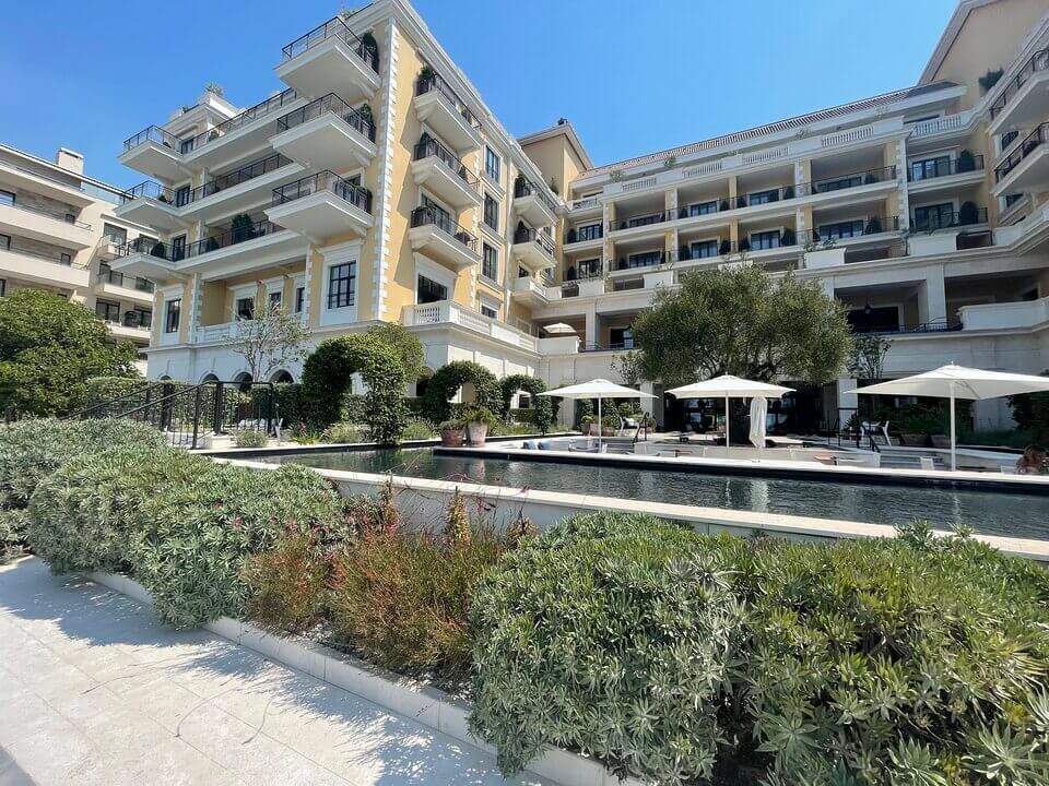 Ein luxuriöses Hotel mit mehreren Balkonen, umrahmt von üppigem Grün und einem langen, reflektierenden Pool im Vordergrund, unter einem strahlend blauen Himmel.