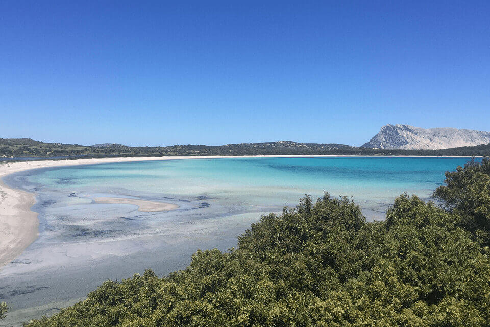 Ein ruhiger Blick auf eine Küstenlandschaft mit kristallklarem, türkisfarbenem Wasser, einem Sandstrand, der sich entlang der Bucht schlängelt, üppigem Grün im Vordergrund und einem markanten Berg im Hintergrund unter einem klaren, blauen Himmel.