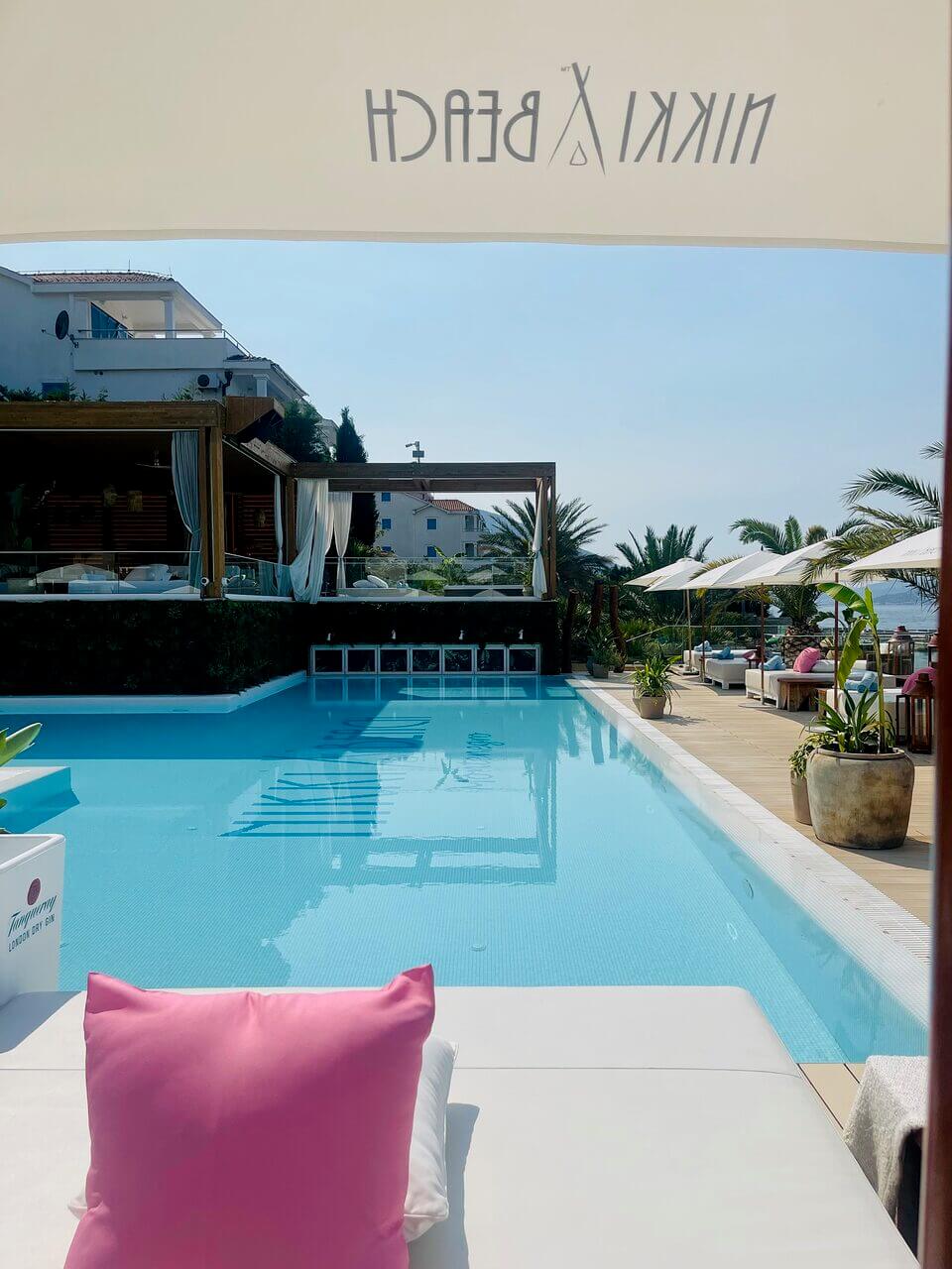 Eine ruhige Poolumgebung mit einem großen Swimmingpool mit strahlend blauem Wasser, umgeben von Palmen und weißen Liegestühlen. Die Aussicht wird teilweise durch eine Markise mit gespiegeltem Text, wahrscheinlich einem Markennamen, verdeckt.