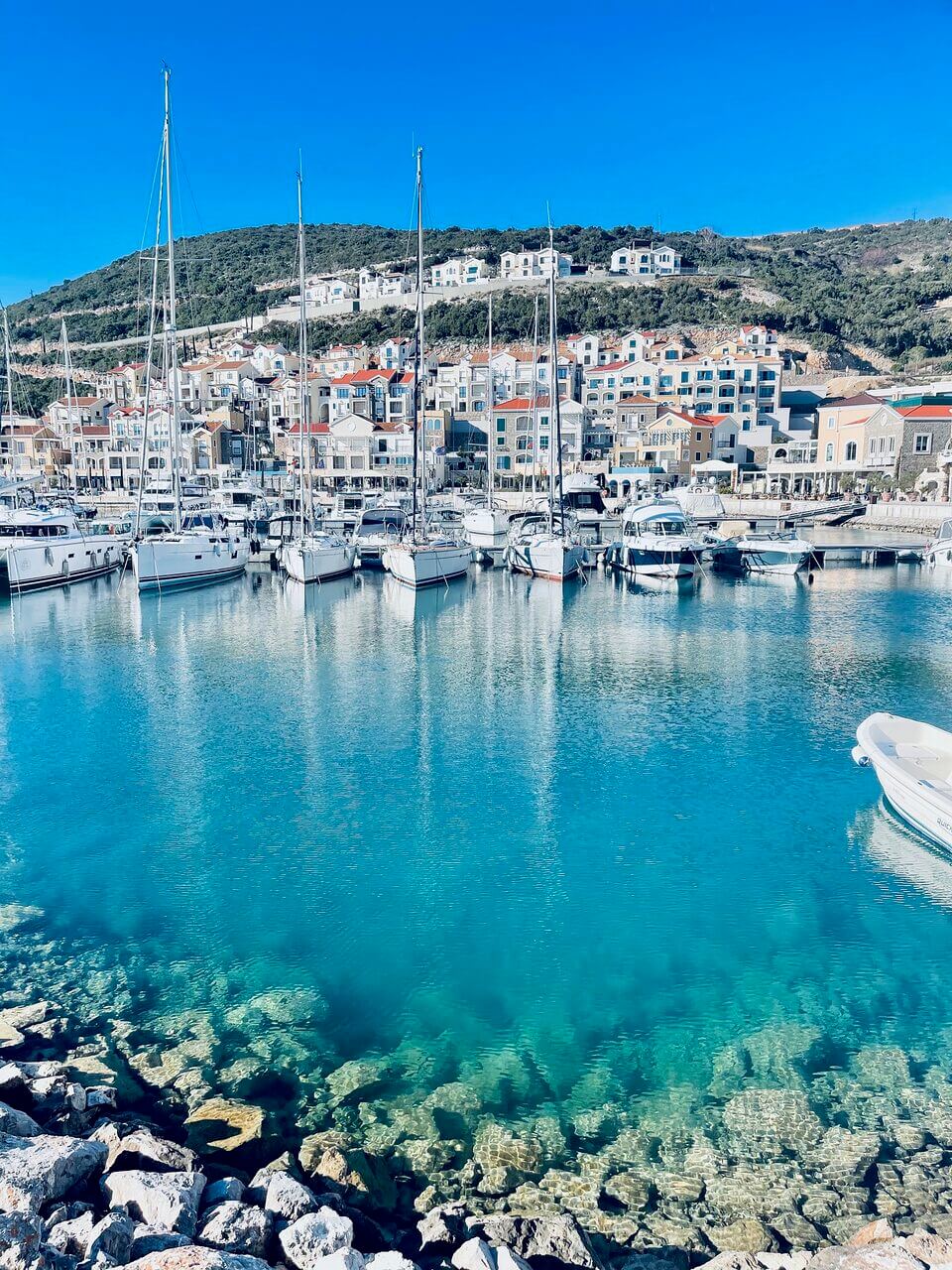 Eine lebendige Küstenszene mit einem Hafen, in dem mehrere weiße Yachten im klaren türkisfarbenen Wasser vor Anker liegen. Im Hintergrund säumen farbenfrohe Gebäude in unterschiedlichen Architekturstilen den Hang unter einem strahlend blauen Himmel.