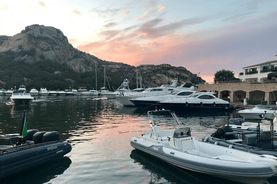 Der Yachthafen Poltu Quato in der Abenddämmerung mit verschiedenen Booten, darunter Yachten und kleinere Motorboote, vor einer Kulisse aus felsigen Hügeln und einem rosafarbenen Himmel. Auf der rechten Seite ist ein Gebäude im mediterranen Stil zu sehen.