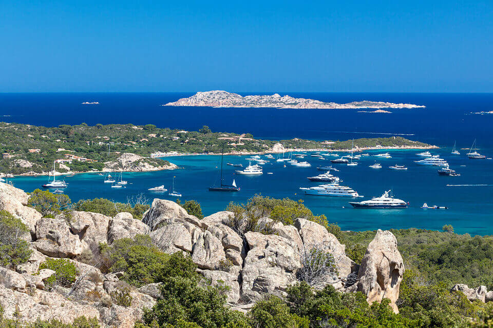 Üppige Küstenlandschaft in der Nähe von Porto Cervo mit klarem, blauem Meer, übersät mit Yachten und Segelbooten in der Nähe eines kleinen Hafens. Felsiges Gelände mit grünem Gebüsch im Vordergrund und eine entfernte Insel unter einem strahlend blauen Himmel.