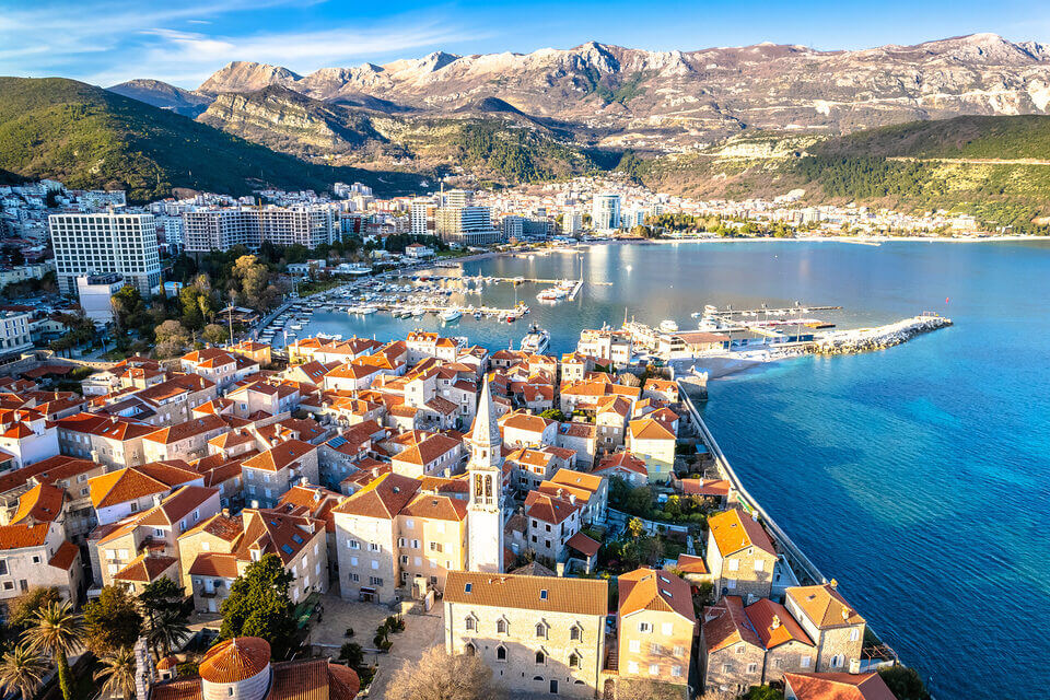 Luftaufnahme des Tourismus in Budva, Montenegro, mit Schwerpunkt auf der Altstadt mit traditionellen Gebäuden mit roten Dächern und einer Kirche, einem modernen Stadtbild, einem Jachthafen mit Booten und den umliegenden Bergen unter klarem blauen Himmel.