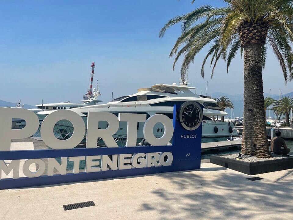 Ein großes Schild „PORTO MONTENEGRO“ mit kreisförmigen Ausschnitten, dahinter sind in einem Yachthafen vertäute Yachten zu sehen, neben einer Palme unter einem klaren Himmel in Tivat.