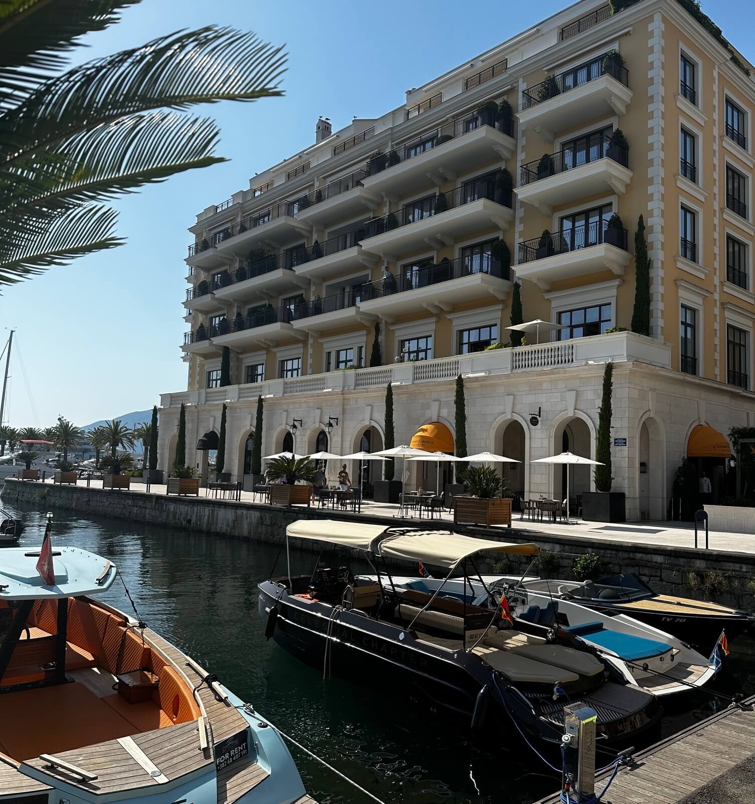Luxuriöses Hotel am Wasser in Montenegro mit mehreren Balkonen mit Blick auf einen Yachthafen mit kleinen Booten und Yachten. Palmen verleihen der ruhigen Umgebung unter einem klaren blauen Himmel eine tropische Note.