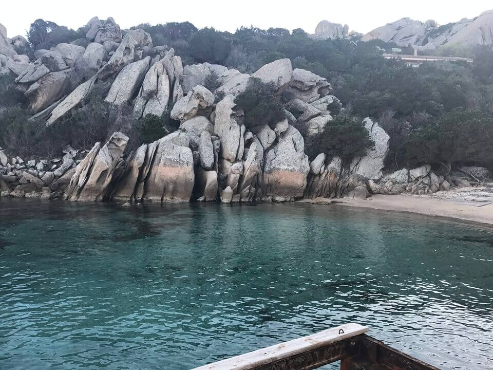 Eine ruhige Küstenszene in Porto Rafael mit klarem Wasser im Vordergrund und markanten, schroffen Felsen, die sich am Wasserrand treffen. Auf einigen Felsen ist Vegetation zu sehen, unter einem bewölkten Himmel. Unten ist die Ecke eines Holzboots zu sehen.