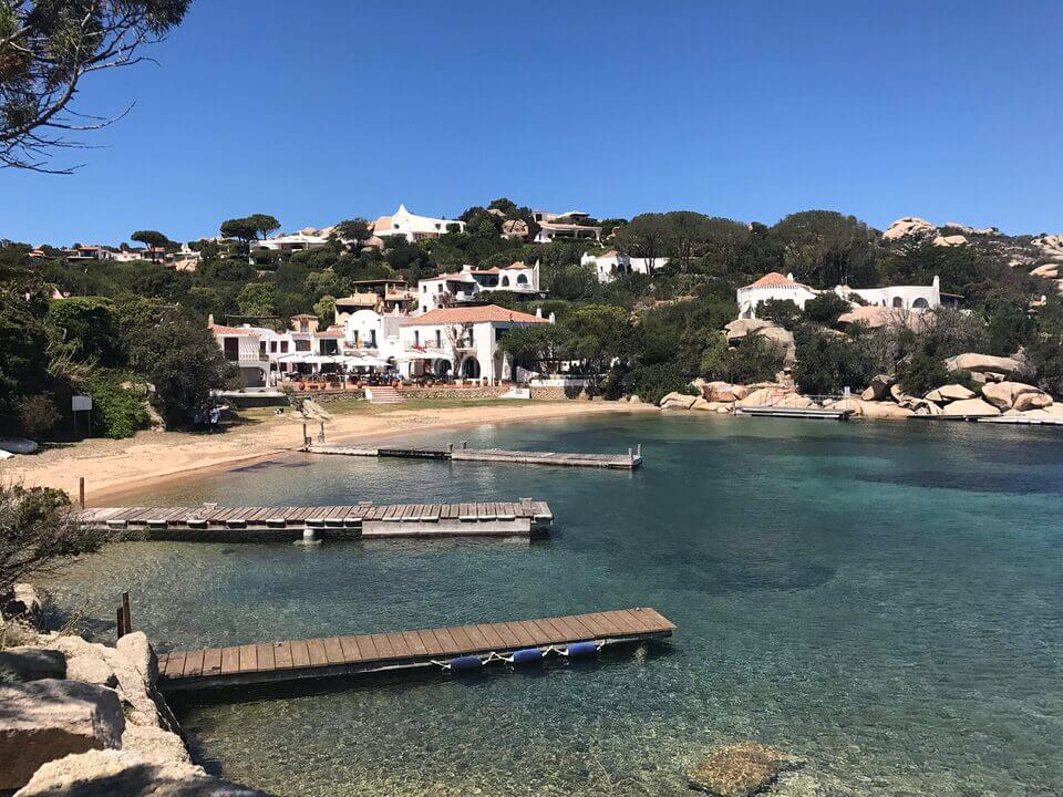 Eine ruhige Küstenszene in Porto Rafael mit kristallklarem türkisfarbenem Wasser und schwimmenden Holzstegen, umgeben von felsigen Ufern und üppigem Grün. Im Hintergrund schmiegen sich weiße Villen im mediterranen Stil unter einen klaren blauen Himmel.