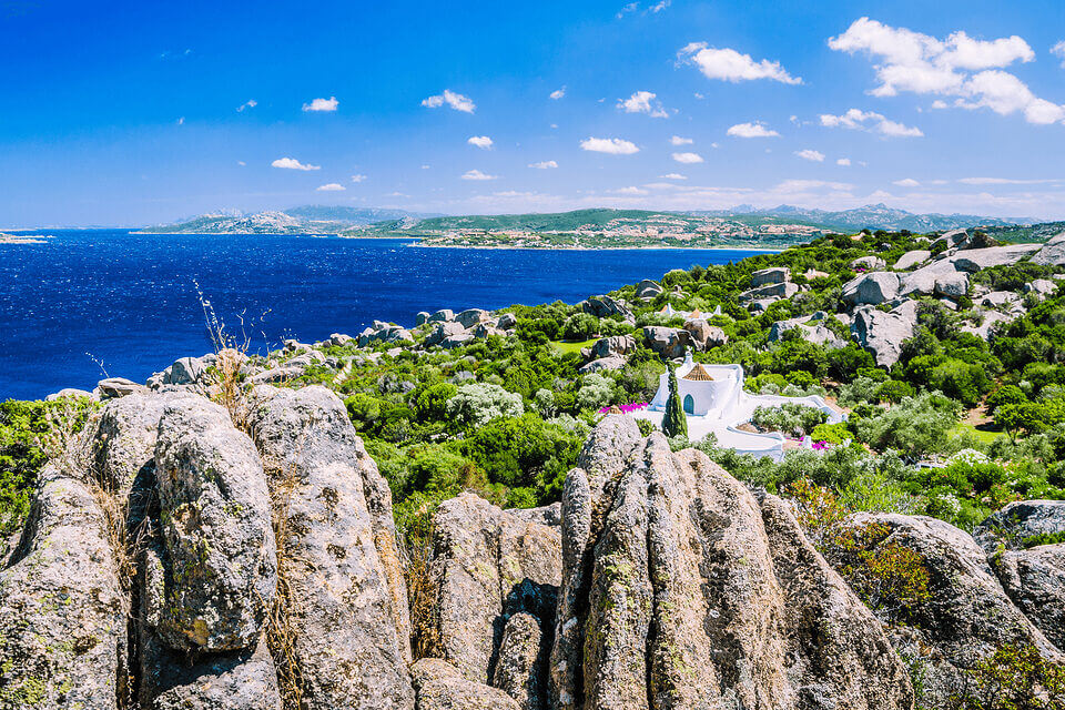 Ein Panoramablick auf eine felsige Mittelmeerküste mit einer kleinen, weißen Kirche inmitten üppigen Grüns, darüber ein klarer blauer Himmel und in der Ferne Hügel jenseits des glitzernden Meeres in Porto Rafael.