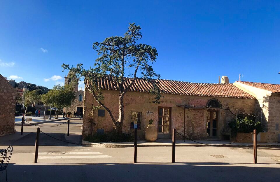 Ein malerisches Steingebäude im Dorf San Pantaleo mit einem Dach aus Terrakottaziegeln unter einem klaren blauen Himmel. In der Nähe wächst ein Baum. Der Bereich ist mit Kopfsteinpflaster ausgelegt, mit rustikalen Geländern und einem Laternenpfahl im Vordergrund.