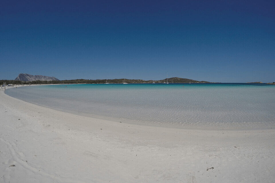 Ein Panoramablick auf die Sandbucht Cala Brandinchi mit puderweißem Sand und kristallklarem türkisfarbenem Wasser. In der Ferne ragen Berge unter einem klaren blauen Himmel auf, und ein einsames Segelboot liegt in Ufernähe vor Anker.