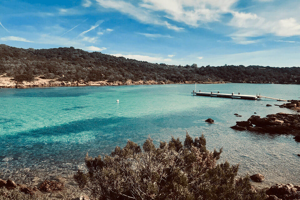 Eine ruhige Küstenszene an der Costa Smeralda mit kristallklarem türkisfarbenem Wasser in der Nähe einer üppig grünen Küste. Ein schwimmender Holzsteg ragt unter einem strahlend blauen Himmel mit schwachen Wolken ins Meer.