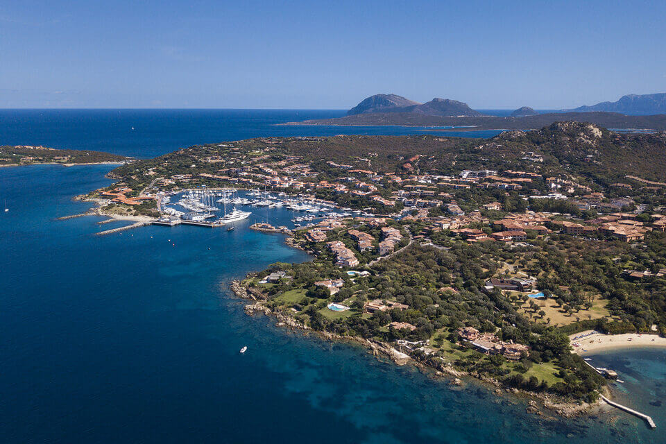 Luftaufnahme von Porto Rotondo, einem Küstendorf mit einem Yachthafen voller Boote, umgeben von klarem, blauem Wasser. Die Landschaft besteht aus verstreuten Gebäuden mit roten Dächern und üppigem Grün, mit Bergen in der Ferne.