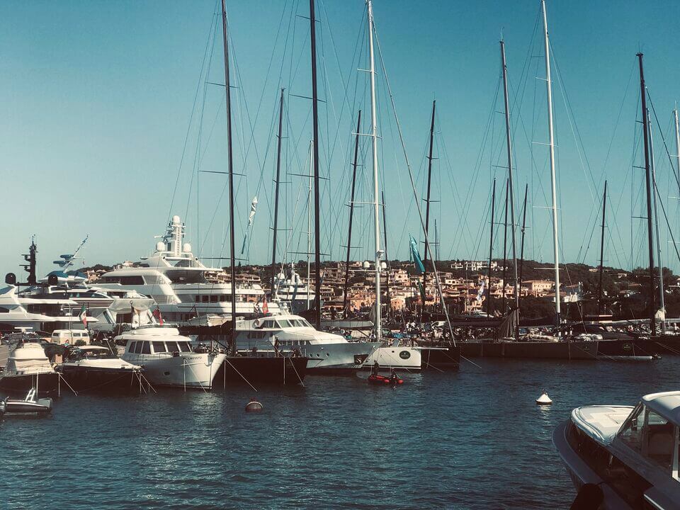 Ein ruhiger Yachthafen in Porto Rotondo, vollgepackt mit verschiedenen Luxusyachten und Segelbooten, deren Masten hoch in die Höhe ragen. Im Hintergrund erhebt sich unter einem klaren blauen Himmel eine Küstenstadt auf einem Hügel.